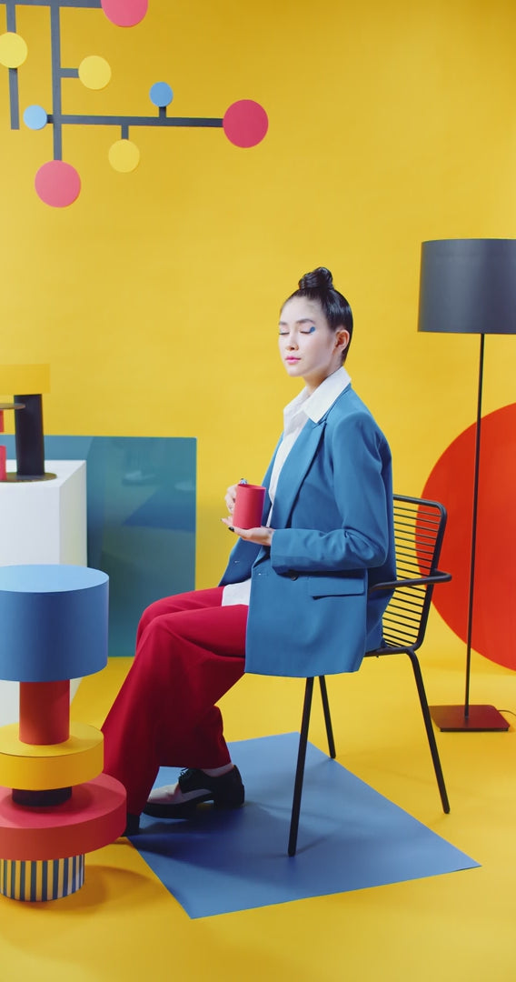 Woman in a blue and red suit in a bright yellow room with abstract, modernist pieces of furniture. She slowly drinks coffee from a bright red cylindrical mug.
